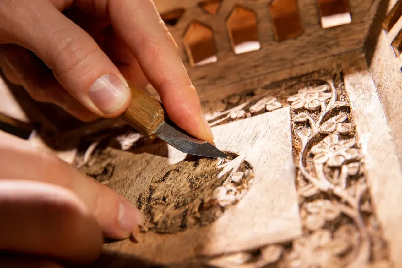 Main d'artisan sculptant un d&eacute;tail floral sur un meuble en bois, &eacute;tape cl&eacute; pour savoir comment nettoyer un meuble en marqueterie.