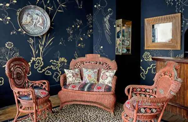 Salon au style napoléon 3, avec des fauteuils et un canapé en rotin corail, sur un tapis léopard, devant un papier peint bleu nuit orné de motifs dorés.