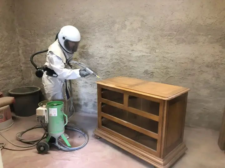 Personne en combinaison intégrale et casque effectuant un décapage bois sur une commode ancienne avec une machine de sablage.